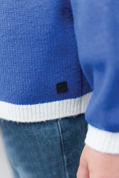 Modro pleten pulover z belimi rebrastimi manšetami in spodnjim robom, z majhno črno tkanino etiketo blizu dna. Približen posnetek teksture in barve.