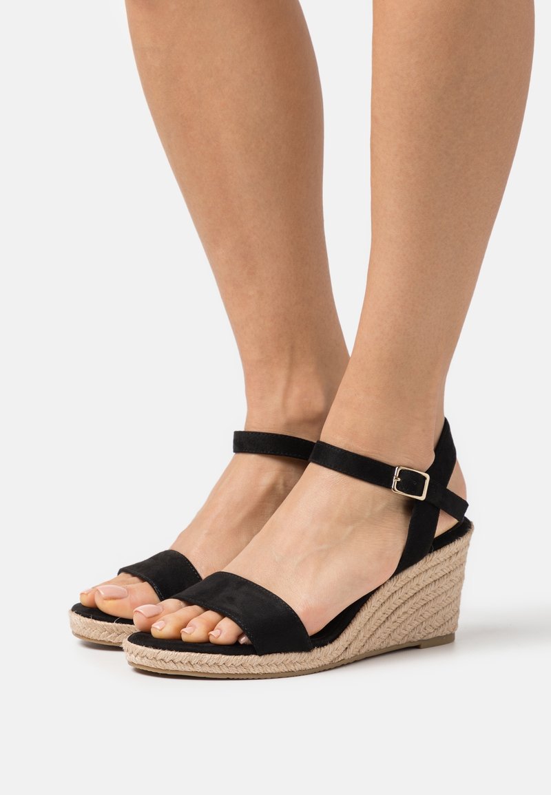 Jambes portant des sandales compensées noires à bride cheville avec une plateforme beige tissée sur fond blanc uni.