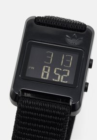 Orologio digitale con quadrante rettangolare nero, che mostra ora e data, e un cinturino in tessuto nero testurizzato. Presenta il logo Adidas in alto.