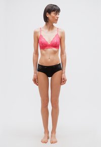 Γυναίκα που στέκεται ξυπόλυτη μπροστά σε λευκό φόντο, φοράει ροζ δαντελένιο σουτιέν και μαύρο δαντελένιο εσώρουχο, κοιτάζοντας προς το πλάι.