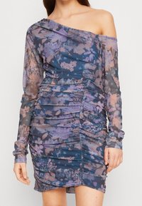 Asymetryczna mini sukienka na jedno ramię z przezroczystego materiału w kwiatowe wzory w odcieniach fioletu i niebieskiego, z marszczeniami po bokach.