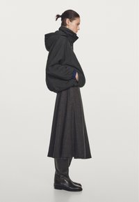 Zwarte gewatteerde jas met een hoge kraag, gedragen over een lange, bruin geruite rok. Zwarte kniehoge laarzen maken de outfit compleet.