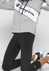 Felpa grigia con un pannello logo in bianco e nero, abbinata a leggings neri. Caratteristiche includono un cappuccio con cordino e un tessuto morbido.