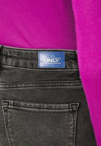 Juodi džinsai iš denim tekstūros, su mėlynu prekių ženklu "Blue Denim ONLY" ant juosmens ir dalinai matoma rožinė viršūnė.