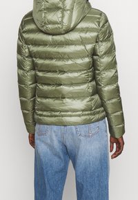 Oliwkowa kurtka puchowa z kapturem, z pikowanym wzorem i błyszczącą fakturą. Noszona z jasnoniebieskimi dżinsami.