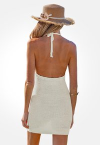 Mujer vistiendo un vestido blanco de punto tipo halter y un sombrero de paja de ala ancha, vista desde atrás sobre un fondo blanco liso.