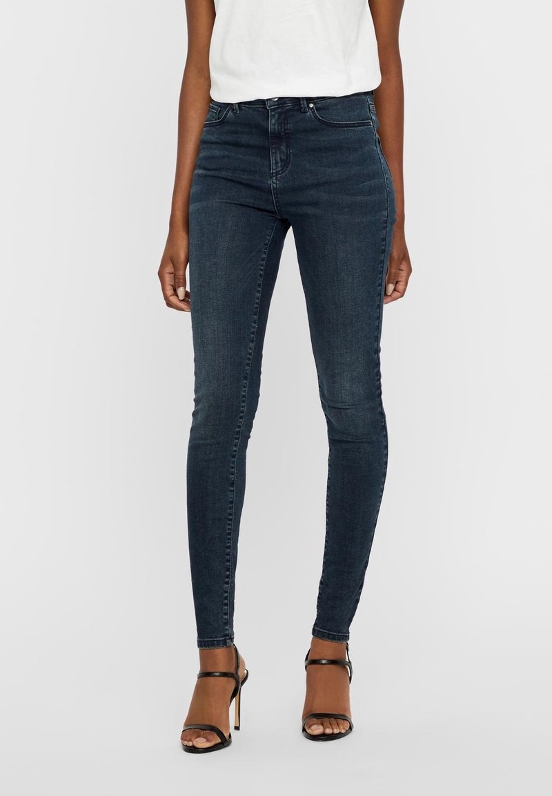 Mörkblå, figurnära jeans i denim; hög midja med subtila sömnadsdetaljer. Kombineras med svarta klackskor med remmar.