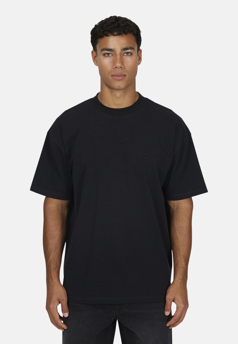 Jeune homme aux cheveux bouclés courts portant un t-shirt noir oversize uni et un jean sombre, se tenant devant un fond clair.