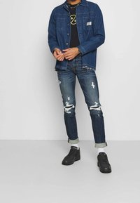 Mörkblå denimjacka för män med knäppning framtill, kombinerad med en svart t-shirt med grafik och ljusblå slitna jeans. Svarta sneakers fulländar looken.