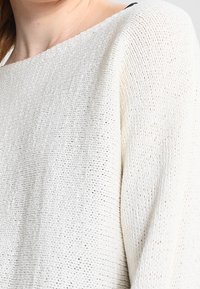 Vit stickad tröja med en lös passform, som har en vid halsringning och textilstruktur. Ärmarna är något överdimensionerade och faller mjukt.