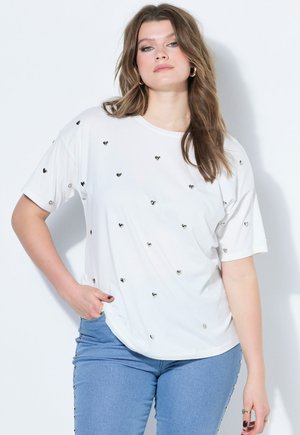 Donna con capelli lunghi castani indossa una maglietta bianca decorata con piccoli cuori d'argento e jeans blu con borchie laterali, posando contro un muro bianco.