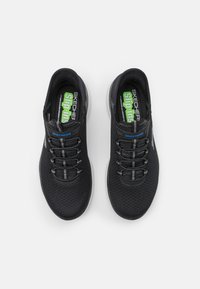 Black slip-on sneakers with a mesh upper, elastic laces, cushioned sole, and branded insole featuring green and blue accents.