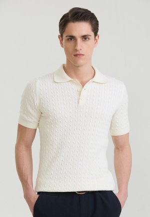 Ung mand med mørkt hår iført en hvid kabelstrikket kortærmet poloskjorte og mørke bukser, der står mod en ensfarvet lys baggrund.