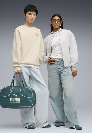 Zwei Models tragen beige und weiße Puma-Oberteile, hellblaue Jeans, türkisfarbene Puma-Schuhe und halten eine türkisfarbene Puma-Reisetasche vor einem grauen Hintergrund.