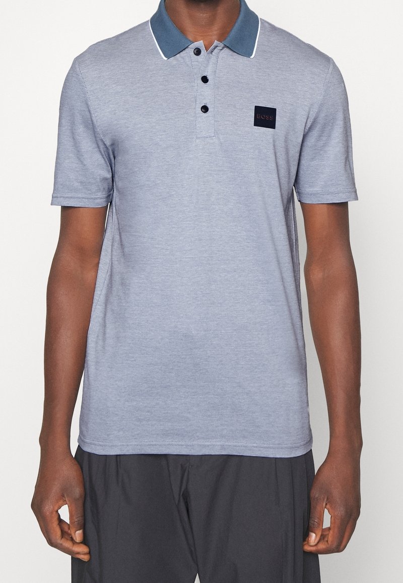Homme portant un polo bleu clair avec un col bleu foncé et trois boutons noirs, associé à un pantalon gris foncé.