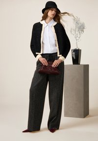 Zwart en crème gebreide cardigan, witte blouse, grijze wijde broek en een kastanjebruine geweven clutch. Model draagt een zwarte hoed.