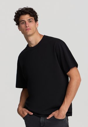 T-shirt nera a maniche corte realizzata in tessuto morbido, con collo a girocollo, vestibilità ampia e design minimalista. Indossata con jeans scuri, mani in tasca.