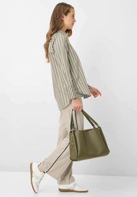 Borsa tote verde oliva con doppio manico superiore, texture liscia e forma strutturata. Maglia a righe a maniche lunghe e pantaloni beige completano il look.