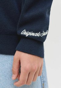 Gros plan sur une main portant une manche tricotée bleu foncé avec le texte brodé en blanc "Originals Studio", et un jean en denim bleu clair.