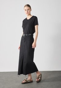 T-shirt noir, associé à une jupe en denim noire taille haute avec une fente avant. porté avec des sandales à glissière ornées de motifs marron et crème.