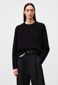 Frau mit glatten braunen Haaren, die einen schwarzen Pullover über einem weißen Oberteil und schwarze, mit einem Gürtel versehene Hose trägt, steht mit einer Hand in der Tasche.