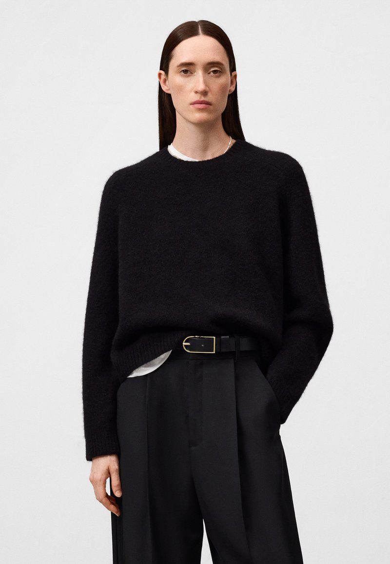 Frau mit glatten braunen Haaren, die einen schwarzen Pullover über einem weißen Oberteil und schwarze, mit einem Gürtel versehene Hose trägt, steht mit einer Hand in der Tasche.