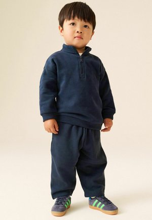 Ungt barn står iført en mørkeblå sweatshirt med lynlås, matchende bukser og grå sneakers med grønne striber på en lys baggrund.