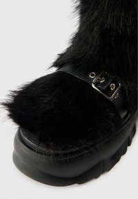 Črne platform čevlje z teksturiranim umetnim krznom, usnjenim trakom s srebrno zaponko in debelim gumijastim podplatom.