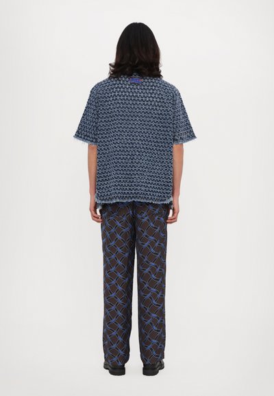 Short-sleeve navy shirt with textured weave and fringe hem, paired with brown patterned pants featuring blue geometric lines. Black shoes.
