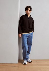 Sort pullover med en kvart lynlås, båret over en beige skjorte. Sammen med lyseblå, løstsiddende jeans og hvide sneakers med grå detaljer.