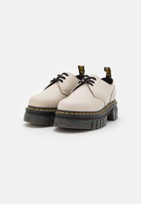 Chaussures beige à lacets en cuir lisse. Semelle en caoutchouc noir épais avec des accents texturés et des coutures jaunes. Deux languettes au niveau du talon.