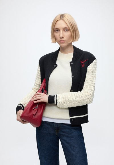 Jeune femme aux cheveux blonds courts portant un pull noir et blanc, un haut blanc, un jean bleu, tenant un sac à main rouge sur un fond uni.