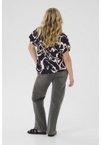 Femme aux longs cheveux blonds, portant un haut à motifs noir et blanc à manches courtes, un jean gris et des sandales noires, se tenant devant un fond uni.