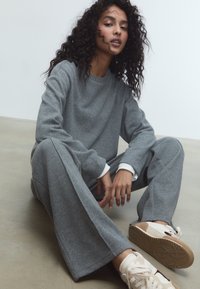 Sivý pletený sveter s dlhým rukávom a široké nohavice s jemnou textúrou. Obsahuje biele akcenty na manžetách. Pohodlné obuv s béžovou podrážkou.