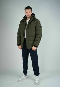 Homme portant une veste matelassée olive avec capuche, une chemise blanche, un jogging bleu marine et des baskets blanches, se tenant contre un fond clair uni.