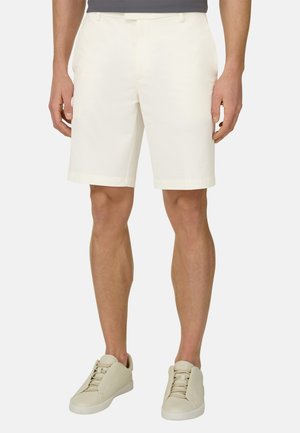 Uomo con pantaloncini bianchi fino al ginocchio e sneakers beige con lacci, in piedi davanti a uno sfondo chiaro uniforme.