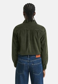 Person with braided hair wearing a long-sleeve olive green shirt and high-waisted blue jeans, standing against a plain background.