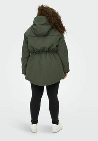 Grön huva jacka med snörd midja, mjuk textur och längre bakstycke, i kombination med svarta leggings och vita sneakers.