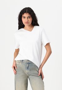 T-shirt bianco a maniche corte con scollo a V e orlo asimmetrico. Abbinato a jeans larghi azzurri, leggermente sbiaditi. Tessuto morbido e casual.