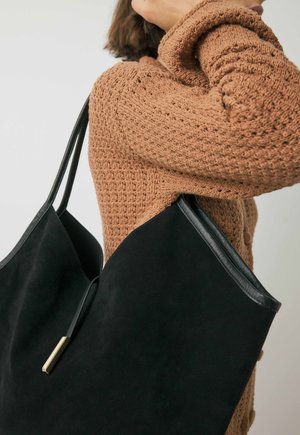 Persona che indossa un maglione di maglia marrone mentre tiene una grande borsa a tracolla in suede nera con spalline sottili e un accento dorato.