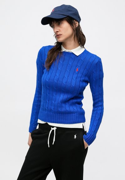 Kék kötött pulóver bordázott szegéllyel, fehér galléros ing fölött viselve. Fekete joggingnadrággal párosítva, melyen fehér húzózsinór és logó található.