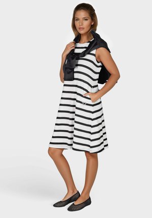 Schwarz-weiß gestreiftes Kleid mit ärmellosem Design, Taschen und einem schwarzen Pullover, der um die Schultern gebunden ist. Kombiniert mit schwarzen Mesh-Ballerinas.