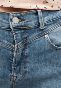 Κοντινό πλάνο μπλε τζιν παντελονιού με κούμπωμα στη μέση, εμφανές κουμπί, θηλιά για ζώνη, τσέπη και πάνω φλοράλ πουκάμισο.