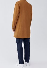 Persoon die met de rug naar de kijker staat, gekleed in een bruin knielang jasje, donkere blauwe cargobroek en witte sneakers tegen een eenvoudige achtergrond.