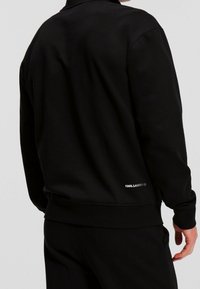 Sudadera negra de material suave con puños y dobladillo acanalados. Presenta un pequeño logo blanco en la parte baja de la espalda. Diseño sencillo, cuello redondo.