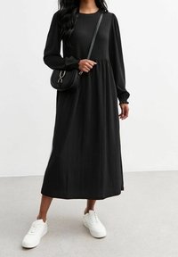 Schwarzes Midikleid mit langen Ärmeln, strukturiertem Stoff und gerafftem Taillenbereich. Kombiniert mit einer Umhängetasche und weißen Sneakers.