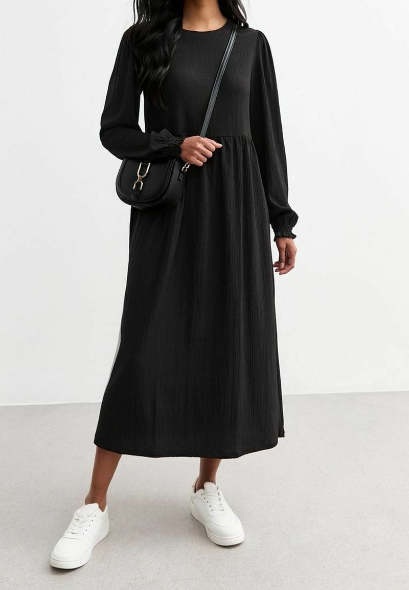 Schwarzes Midikleid mit langen Ärmeln, strukturiertem Stoff und gerafftem Taillenbereich. Kombiniert mit einer Umhängetasche und weißen Sneakers.