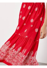 Rotes Kleid aus leichtem Stoff, mit weißen dekorativen Mustern und einem gerafften Saum sowie einem geschichteten, fließenden Design.