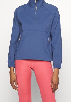 Niebieska bluza z suwakiem do ćwiartki i bocznymi kieszeniami na zamek, zestawiona z jasnoróżowymi legginsami, na stojącej osobie na białym tle.