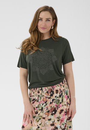 Donna con capelli castani lunghi indossa una maglietta verde scuro con motivo floreale a puntini e una gonna con disegno floreale colorato su sfondo semplice.